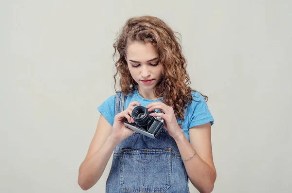 Bruna riccia in tuta di jeans tiene una macchina fotografica tra le mani, torce le leve . — Foto Stock