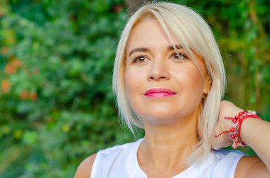 Yeşil yapraklı güzel sarışın, orta yaşlı kadın. Beyaz bluzlu, caddede duruyor. Güzel bir gülümseme düz kısa saçlar, kahverengi gözler