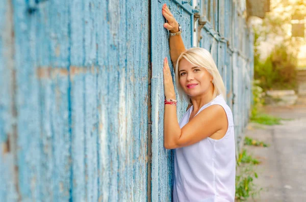 Όμορφη ξανθιά γυναίκα σε μια λευκή μπλούζα αγκαλιασμένη σε ένα παλιό μπλε ξύλινο τοίχο, τα χέρια ψηλά. Ένα όμορφο χαμόγελο στο πρόσωπο ίσια κοντά μαλλιά, καστανά μάτια — Φωτογραφία Αρχείου