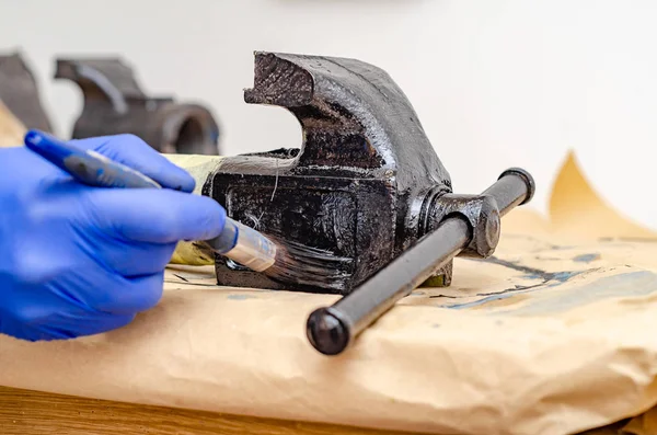 Hand in blue rubber gloves paints metal parts of an old vise with black paint with a brush — Stock Photo, Image