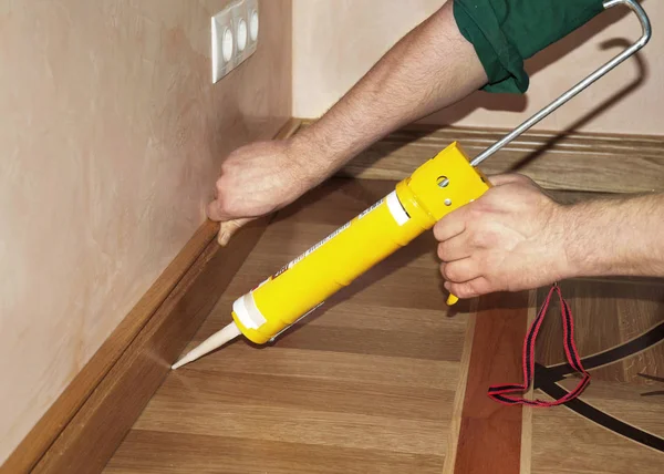 Riparatore che installa il pavimento di legno della quercia del battiscopa con la colla della pistola di calafataggio dalla cartuccia . — Foto Stock