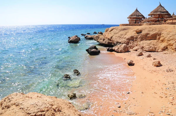 Ombrelloni in spiaggia con ombrelloni in paglia e lettini in legno davanti ad un turchese sulla spiaggia del Mar Rosso con onde e cielo azzurro . — Foto Stock