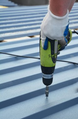 Koruyucu eldivenli bir müteahhit çatıya tornavidayla metal bir kiremit yerleştiriyor..