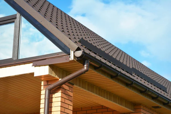 Gable roofing construction with brown metal tiled roof, a rain gutter, snow guards and a skylight against blue sky.