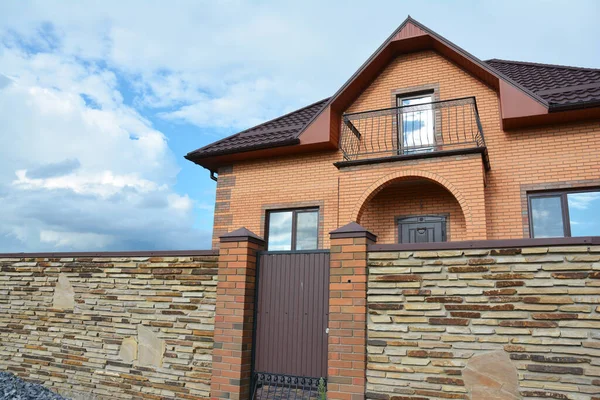 Modern brick house construction with bonnet rooftop covered with metal roof tiles, open balcony and brick fence tiled with faux brick panels agains blue sky.