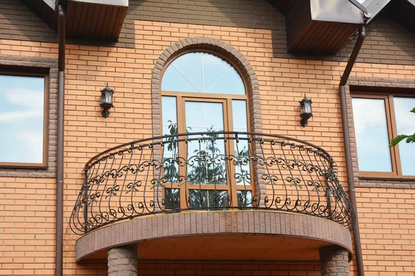 A close-up on semi-circled balcony with wrought iron railings, arched glass door and roof gutter system with downpipes of a brick house.