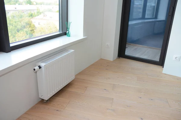 An apartment room with a black profile window and black profile glass balcony door, white radiator heater and wood laminated floor.