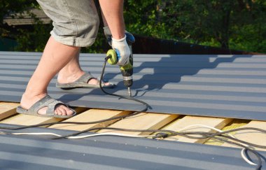 Bir müteahhit, evin çatısına elektrikli tornavida kullanarak kutu profili örtüsü, boya kaplı metal çatı örtüsü kuruyor.. 