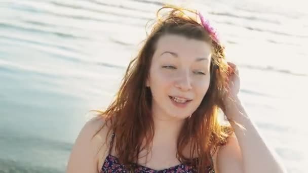 Chica joven feliz con flor en el pelo en la playa sonrisa en la cámara. Día soleado. Mar. — Vídeo de stock