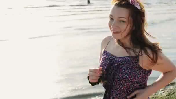 Chica joven feliz con flor en el pelo ejecutar en la playa sonrisa en la cámara. Día soleado . — Vídeo de stock