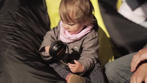 Sint-Petersburg, Rusland-16 juli 2015: Little Bay meisje zit op Zitzak Touch grote koptelefoon. Kindertijd. Zomer winderige dag. Openluchtfestival — Stockvideo