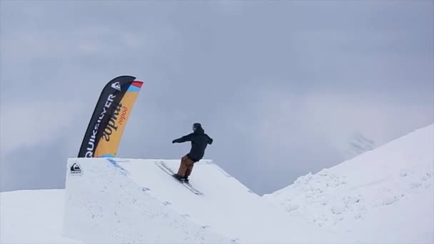 СОЧИ, РОССИЯ - 29 марта 2016 года: Сноубордист в форме прыгает с трамплина, сгибает ноги в воздухе. Снежные горы. Пейзаж — стоковое видео