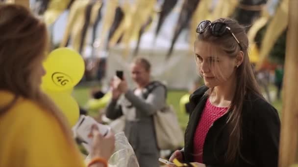 СЕНТ-ПЕТЕРБУРГ, РОССИЯ - 16 июля 2015 года: Девушка подарит женщине белый подарочный бокс, желтые и черные теннисные ракетки, открытки на летнем фестивале. Солнечный день — стоковое видео