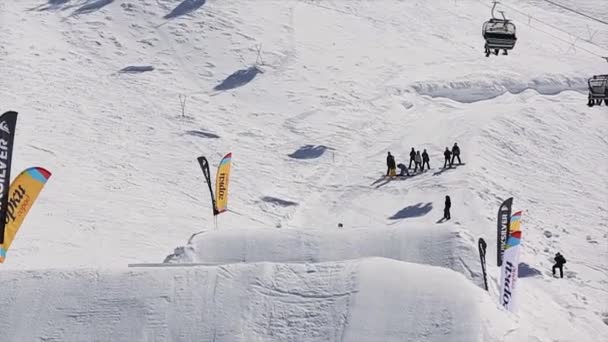 Soči, Rusko - 1. dubna 2016: Snowboardista, skok do výšky z odrazový můstek se převrátit ve vzduchu. Zasněžené hory. Slunečný den. Lidé — Stock video