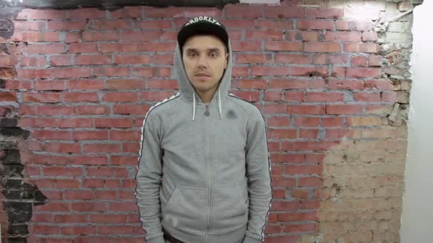 Man in grey hoody, black cap speak something on camera. Brick wall on background — Stock Video