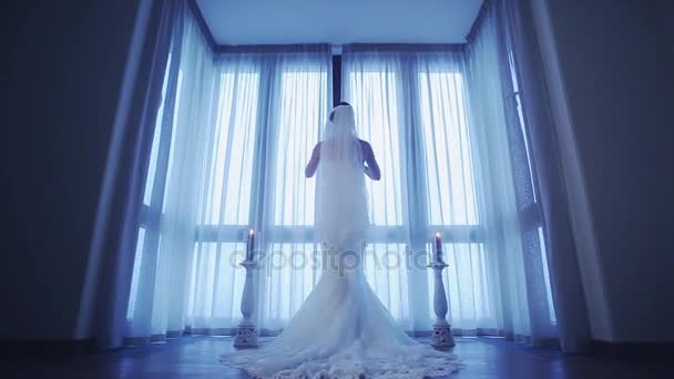 Parte posterior de la novia en vestido de novia vienen a la ventana y destapar cortinas blancas. Sombras frías . — Vídeo de stock