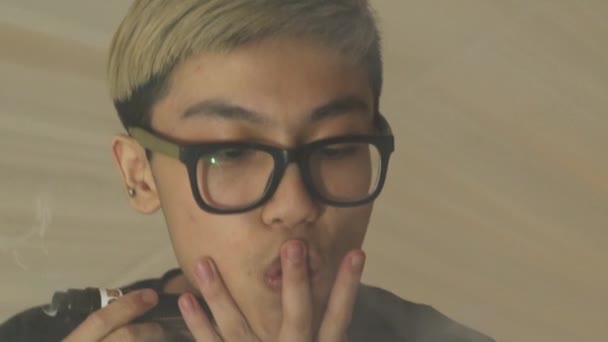 Азиатский мальчик в очках выдыхает двойные круги пара из электронной сигареты. Vape — стоковое видео