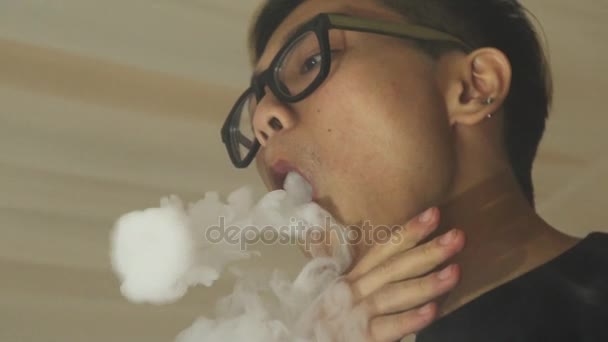 Азиатский мальчик в очках выдыхает кольца из электронной сигареты. Вэйпер. Субкультура — стоковое видео