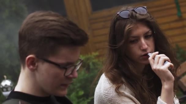 Girl and man in glasses exhalate steam from electronic cigarette on bench. Vaper — Stock Video