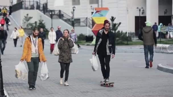 СОЧИ, РОССИЯ - 27 марта 2016 года: Вид трех друзей, идущих по улице с большими белыми пакетами. Один человек на скейтборде. Вечер в городе — стоковое видео
