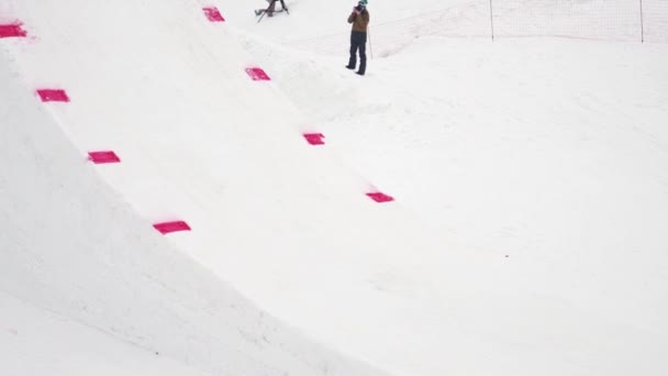 SOCHI, RUSIA - 4 DE ABRIL DE 2016: Paseo de snowboarder en trampolín, hacer voltear en el aire. Montañas nevadas. Clima gris — Vídeos de Stock