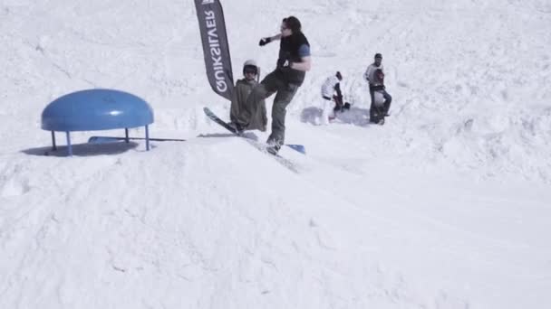 SOCHI, RUSIA - 1 DE ABRIL DE 2016: Estación de esquí. Snowboarder saltar en el trampolín. Camarógrafo. Montañas nevadas. Soleado. — Vídeo de stock