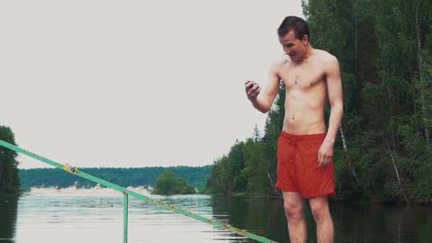 若い男は、手にストップウォッチが付いている湖に立っています。自然。夏の日。感情 — ストック動画
