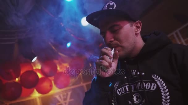 Sankt Petersburg, Ryssland - 28 maj, 2016: Mannen i keps rök elektronisk cigarett på scenen av nattklubb, andas ut ånga. — Stockvideo