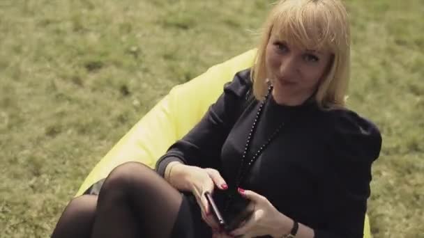 Fille blonde en robe noire assis sur un sac de haricot jaune sur l'herbe verte, posant sur la caméra, sourire. Été — Video