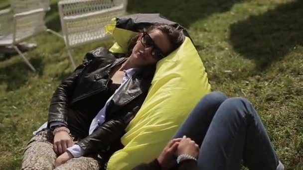 Dos chicas en gafas de sol relajándose en bolsas de frijoles amarillos en hierba verde, sonrisa. Festival de verano. Descanso — Vídeos de Stock