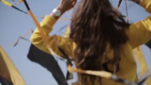 Boldog lány sárga kapucnis napszemüveg emelje fel a kezét között hullámzó szalagok. Nyári szeles napon. Fesztivál — Stock videók