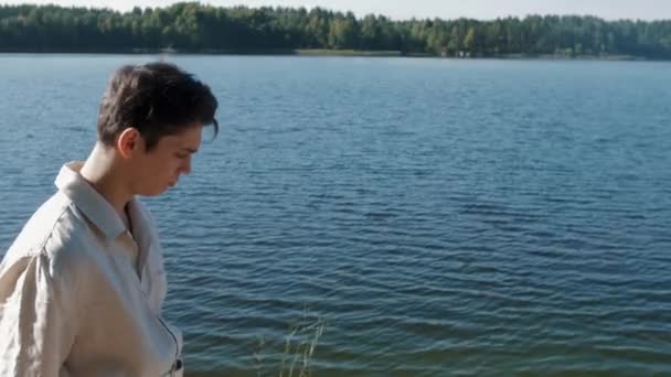 Меланхолик в белой рубашке идет вдоль берега озера в солнечный летний день — стоковое видео