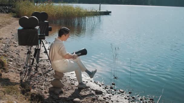Кинорежиссер в сетке для волос ест виноград из динамика на берегу озера с двумя камерами — стоковое видео