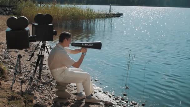 Кинопродюсер в сетке для волос ест виноград из динамика на берегу озера с двумя камерами — стоковое видео