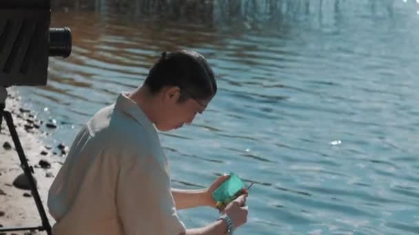 Diretor de cinema em rede de cabelo gritar para alto-falante comer uvas na margem do lago com câmera — Vídeo de Stock