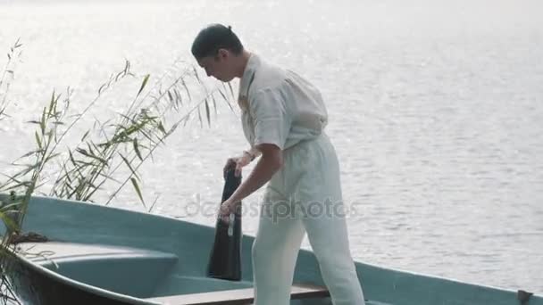 Man in hair net and white clothes standing on boat paddles oar on lake — Stock Video