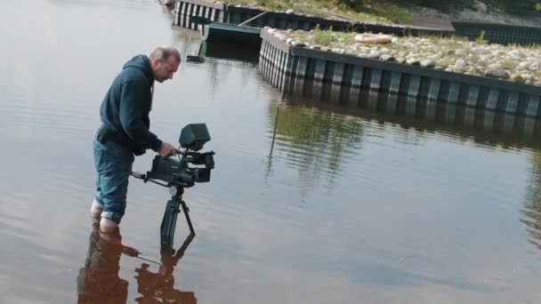 Camarógrafo de pie rodilla profunda en el agua con la cámara en el trípode en verano día soleado — Vídeos de Stock