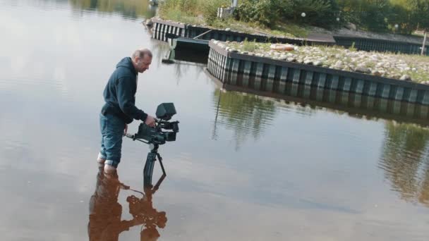 Cameraman permanent knie diep in water met de camera op statief in de buurt van pier zonnige dag — Stockvideo