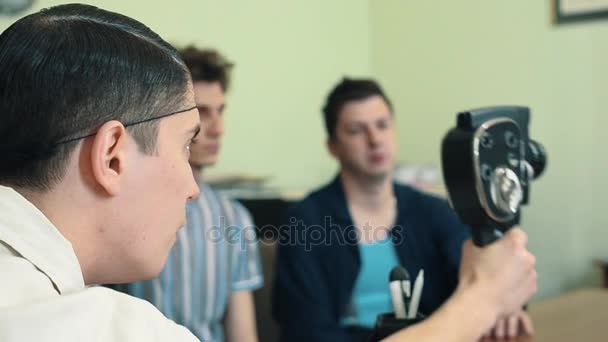 Мужчина в сетке для волос использует старую 8-миллиметровую камеру в офисе, снимая двух рабочих — стоковое видео