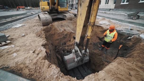 SAINT PETERSBURG, RUSSIA - SEPTEMBER 26, 2016: Worker in hard hat gestures excavator scooping soil in trench at building site — Stock Video