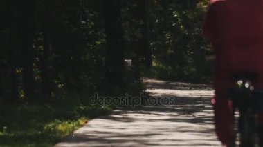Adam kırmızı spor kıyafetli bir ormandaki bir asfalt yol boyunca bir bisiklet ile gidiyor