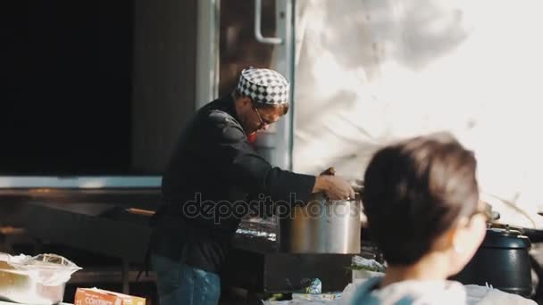 Sint-Petersburg, Rusland - 28 augustus 2016: Cooking chef-kok in GLB en en zwarte chef-kok jas zet grote pot in een brazier buitenshuis — Stockvideo