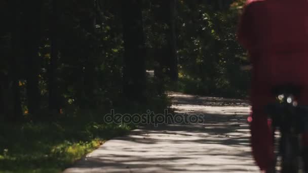Un bărbat în costum de sport roșu merge cu bicicleta de-a lungul unui drum de asfalt într-o pădure — Videoclip de stoc