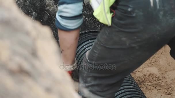 Рабочий втыкает черную пластиковую трубу в круглое отверстие в бетоне в песчаной канаве — стоковое видео