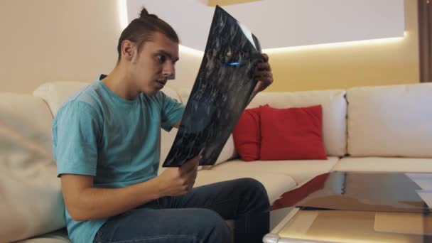 Oroliga killen i blå skjorta sitter på läder soffa titt på tryckta röntgen ark — Stockvideo