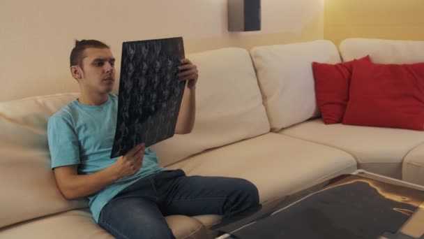 Hombre preocupado en camisa azul sentado en sofá de cuero observa impreso hoja de rayos X — Vídeos de Stock