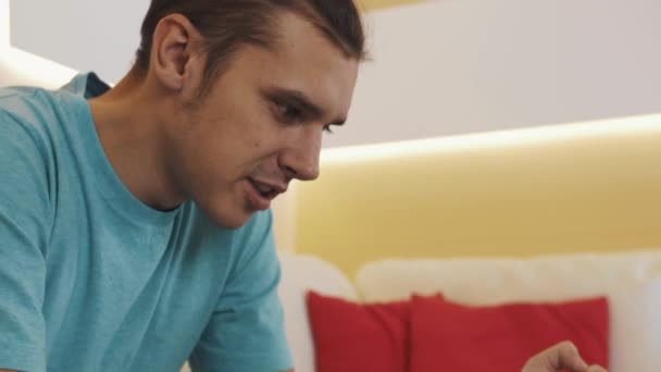 Мужчина в синей рубашке, сидящий на диване, злится, проваливая видеоигру на закрытом планшете — стоковое видео