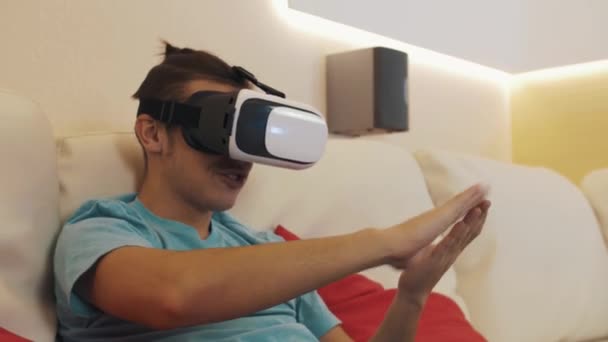 Young man sitting on sofa wears virtual reality headset makes money tossing gesture — Stock Video