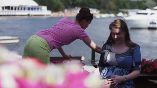 Flicka i rosa skjorta talar till unga gravid kvinna. Sitter på terrassen vid floden. — Stockvideo