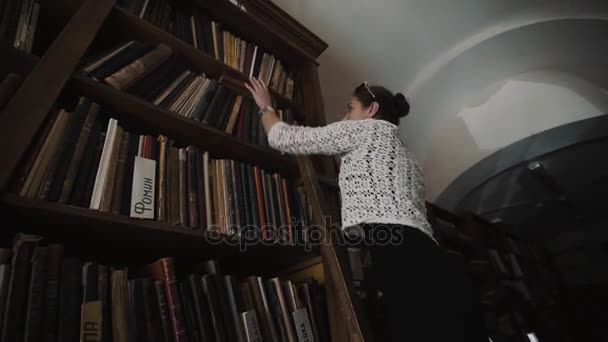 Woman standing on ladder taking book of bookshelf in old style library interior — Stock Video